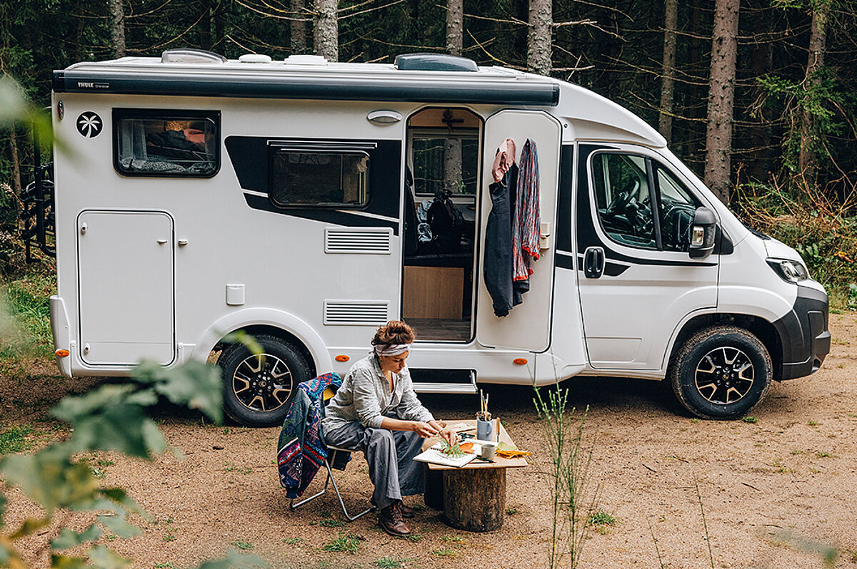 Eine Frau isst vor ihrem Wohnmobil von Sunlight auf einer Waldlichtung.