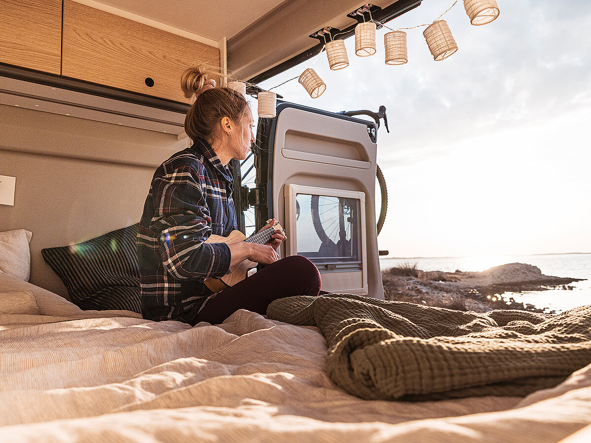 Eine junge Frau spielt Gitarre auf dem Bett eines Kastenwagens, die Tür ist geöffnet und sie schaut raus.