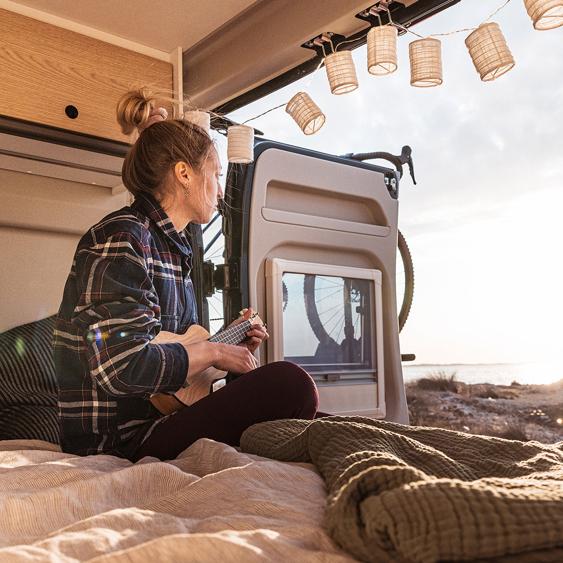 Eine junge Frau spielt Gitarre auf dem Bett eines Kastenwagens, die Tür ist geöffnet und sie schaut raus. 