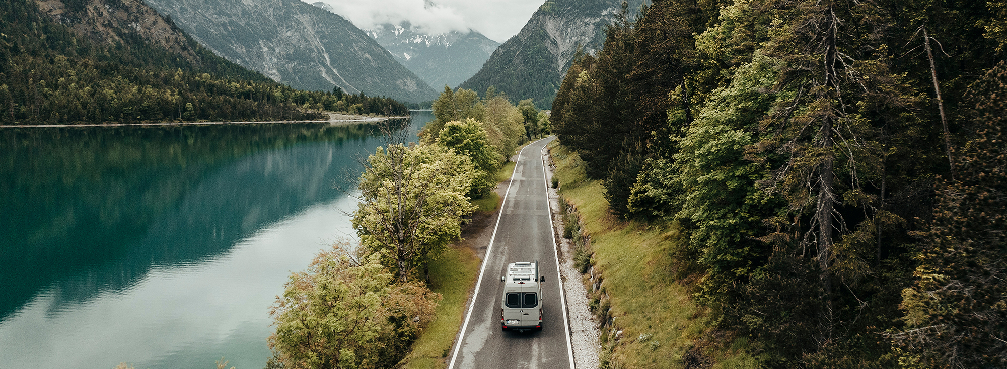 Ein Camper Van fährt eine Landstraße am See entlang.